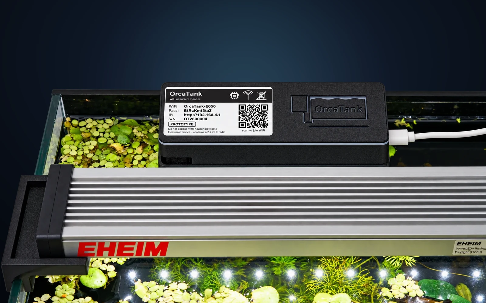 The OrcaTank main unit clipped over the rim of a planted aquarium, beside the light bar, above floating plants